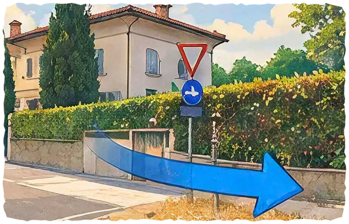 Incrocio davanti a una villa color crema con siepe e cartello di dare precedenza; una grande freccia blu indica la svolta stretta a destra.